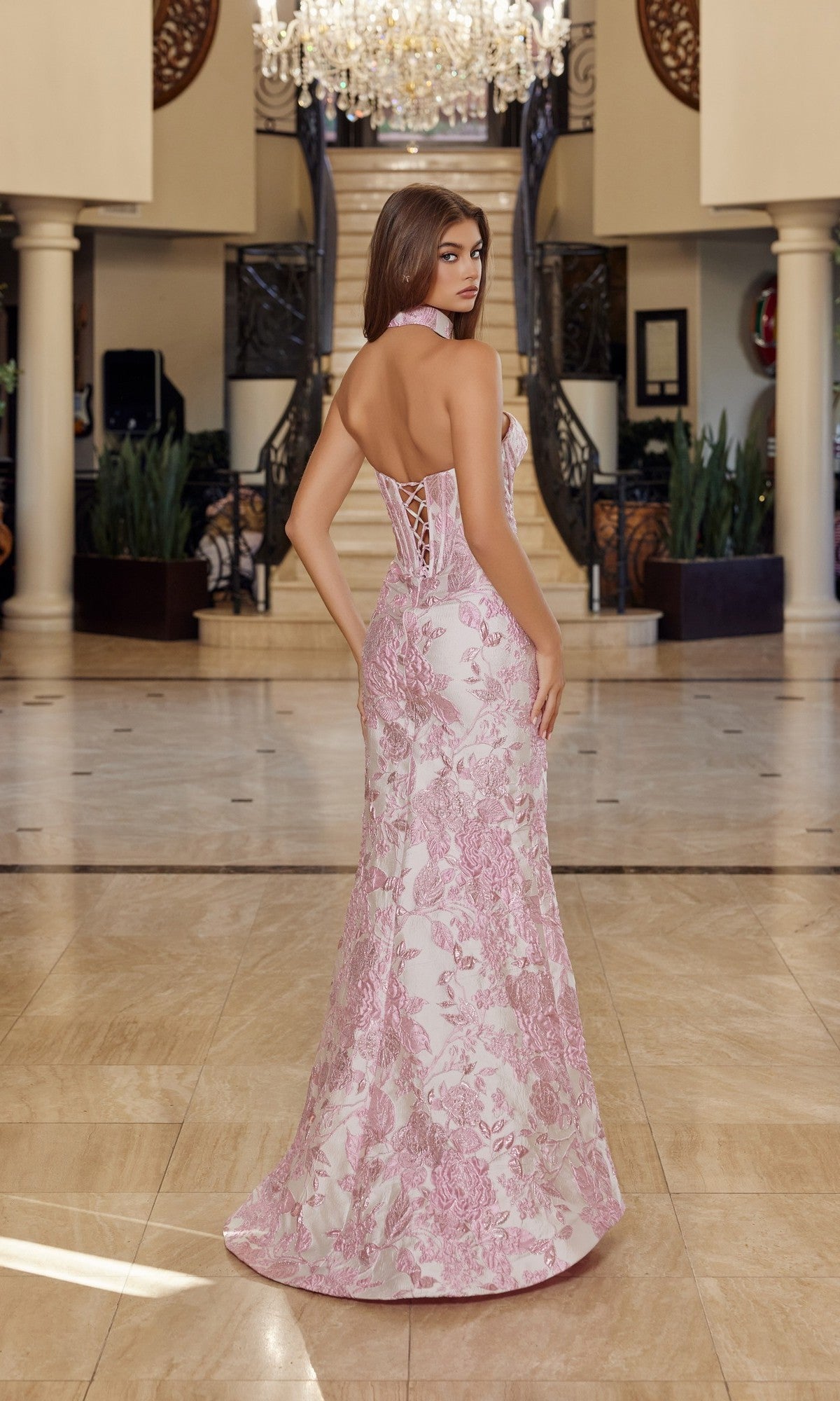A woman stands indoors on marble flooring, facing away, wearing the Nox Anabel T1922 long pink and white floral brocade prom dress with a corset bodice. A grand staircase and chandelier are in the background.